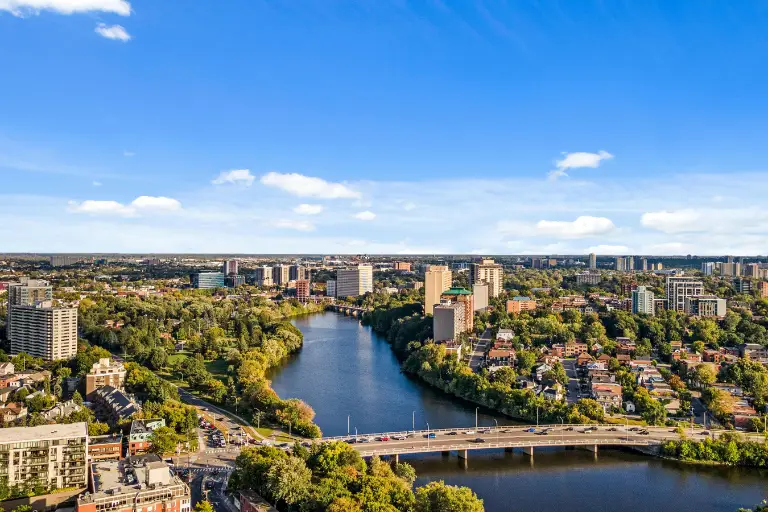An aerial landscape view of Ottawa's green neighbourhoods, residential areas, and a river flowing under a bridge, highlighting the best family communities.