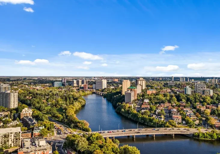 An aerial landscape view of Ottawa's green neighbourhoods, residential areas, and a river flowing under a bridge, highlighting the best family communities.