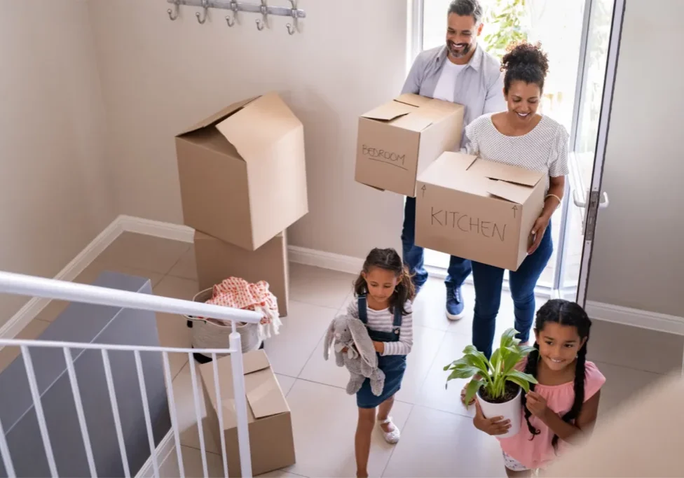 A happy family carrying moving boxes labeled "kitchen" and "bedroom" into their new home in Ottawa, illustrating the guide for first-time home buyers.